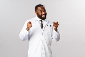 Portrait of friendly handsome doctor pointing at camera, inviting people for check-up, telling stay home be safe during corona, covid19 pandemic, smiling encourage vaccinate, grey background photo