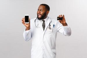 Covid19, pandemic and online medicine concept. Complaining angry african-american man looking at mobile phone display, hold credit card, outraged with huge bills and expensive app subscribtion photo