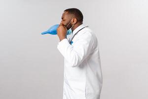 Covid-19, coronavirus patient treatment and disease concept. Funny young african-american male doctor fool around, having break from work, blowing air into latex glove, playing have fun photo