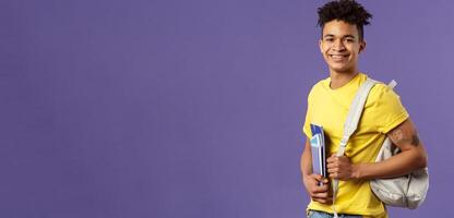 Back to school, university concept. Portrait of handsome charismatic hispanic guy, student walking to campus, going for lecture, studying hold backpack and notebooks, stand purple background photo