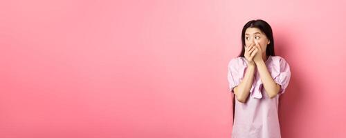 Shocked and startled asian woman looking aside at logo, covering mouth with hands speechless, standing in dress on pink background photo