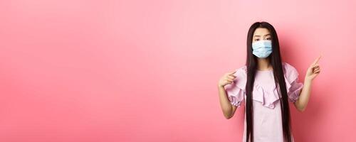 Healthy people and covid-19 pandemic concept. Bored asian woman in medical mask pointing right, showing logo, standing unamused on pink background photo