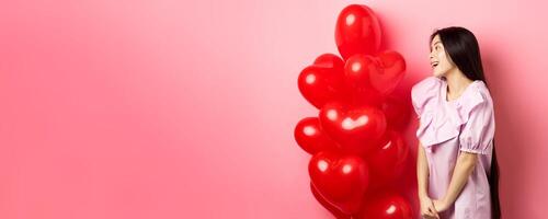 Romance and love concept. Romantic asian girl looking with affection and sympathy at empty space, standing near red hearts balloons from lover, pink background photo