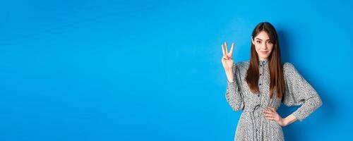 Young beautiful girl in dress with natural long hair, showing number three with fingers and smiling, standing against blue background photo