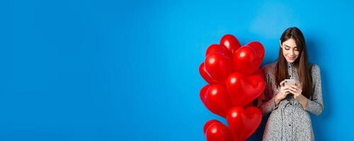 Valentines day. Smiling woman in dress standing near red hearts balloons and looking at smartphone, standing on blue background photo