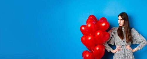 Valentines day. Annoyed and bothered girl complaining, looking left at empty space and frowning, standing near heart balloons, blue background photo
