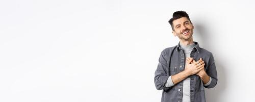 Romantic guy hold hands on heart and smiling, thanking you, looking pleased and grateful, standing on white background photo