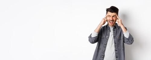 Distressed young man touching head and shouting, looking frustrated aside, standing on white background photo