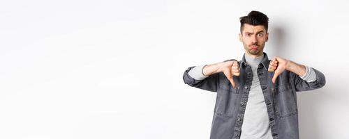 Disappointed funny man frowning and looking upset, showing thumbs down displeased, standing on white background photo