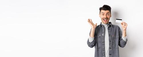 Excited man scream from joy and showing plastic credit card, rejoicing on shopping, standing against white background photo