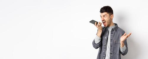 Angry guy shouting at speakerphone, scream on mobile phone, leaving angry voice message, standing against white background photo