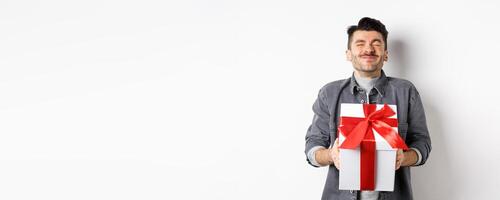 Happy handsome young man holding big valentines day gift and cheering, receive romantic surprise present from lover, standing on white background photo
