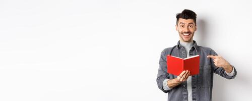 Happy smiling guy pointing at red planner, showing good journal or book, standing on white background photo