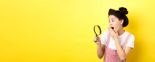 Excited silly asian girl gasping wondered, found something interesting, looking through magnifying glass, standing on yellow background photo