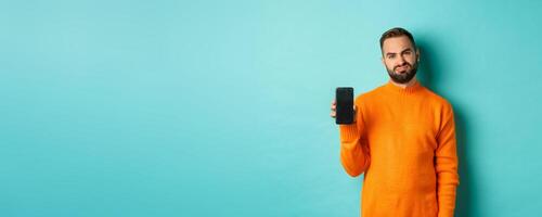 Displeased and disappointed man in orange sweater, grimacing and showing mobile screen, standing over light blue background photo