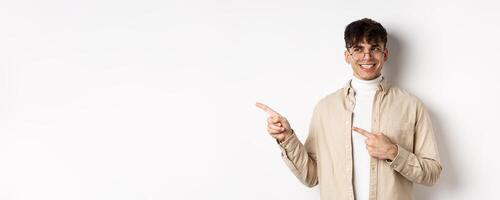 Handsome male model in glasses and trendy clothes, looking and pointing left at empty space, smiling with happy and pleased face expression, white background photo
