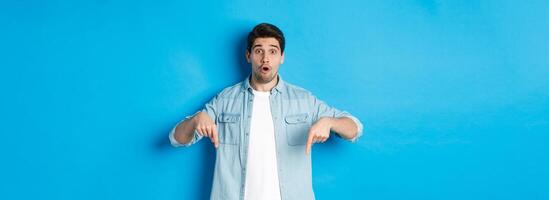 Surprised bearded adult man in casual outfit, pointing fingers down and looking startled, asking question about promotion, standing over blue background photo