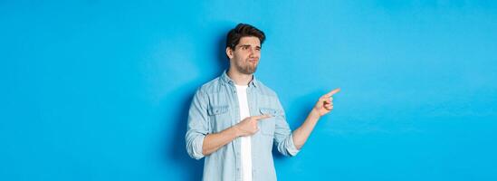 Doubtful adult man pointing fingers left at promotion and looking unsure, grimacing disappointed, standing against blue background photo