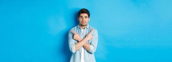 Portrait of nervous guy pointing fingers sideways, looking indecisive and asking for help with choice, showing left and right promo offers, standing against blue background photo