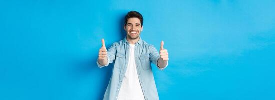 Smiling confident man showing thumbs up, guarantee quality, approving something good, standing against blue background photo