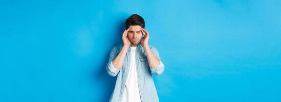 Man with hangover touching head and grimacing, having headache, standing against blue background photo