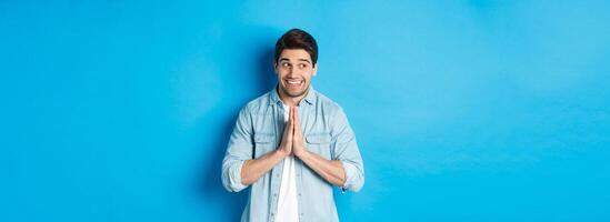 Hopeful smiling man looking left, holding hands in pray, begging for something or pleading, standing against blue background photo
