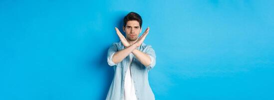 Displeased man frowning and saying no, making cross sign, tell to stop, standing against blue background photo