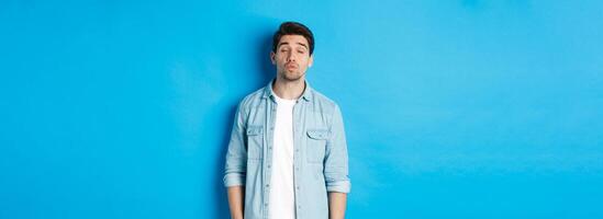 Handsome man waiting for kiss, pucker lips and close eyes while standing against blue background photo