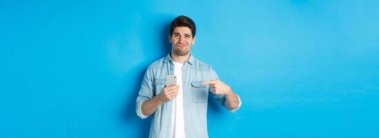 Concept of online shopping, applications and technology. Skeptical and displeased guy pointing finger at smartphone and grimacing disappointed, standing over blue background photo