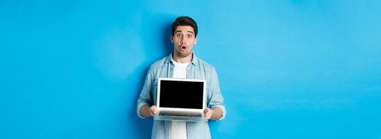 Man showing advertisement on laptop screen and looking amazed, saying wow and looking at camera, standing against blue background photo