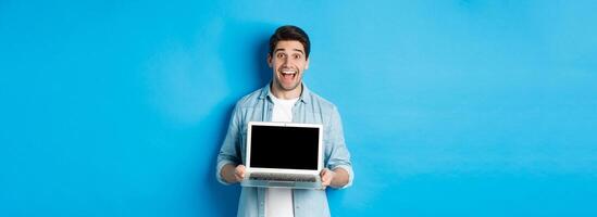 Cheerful smiling man making presentation, showing laptop screen and looking happy, standing over blue background photo