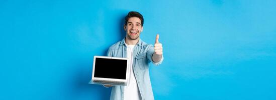 Image of satisfied handsome man showing laptop screen, thumbs-up in approval, like website or internet, standing over blue background photo