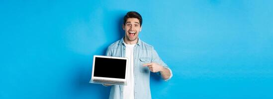 Excited smiling man pointing at laptop screen, showing internet promo offer or website advertisement, standing over blue background photo