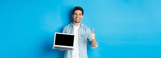 Smiling handsome man showing laptop screen and thumb-up, like promo offer, recommending website, standing against blue background photo