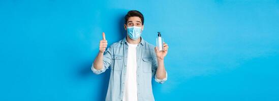 Concept of covid-19, pandemic and social distancing. Cheerful man in medical mask, showing hand sanitizer and thumbs-up, recommending sanitizer, standing against blue background photo