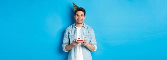 Smiling man holding b-day cake and wearing birthday party hat, celebrating over blue background photo