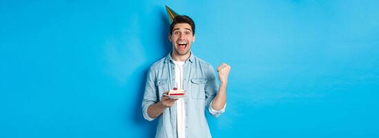 Cheerful man looking happy, celebrating birthday in party hat, holding b-day cake and make fist pump, standing over blue background photo