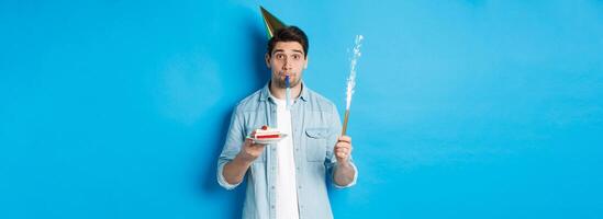 Funny guy celebrating birthday, holding b-day cake, firework and wearing party hat, standing over blue background photo