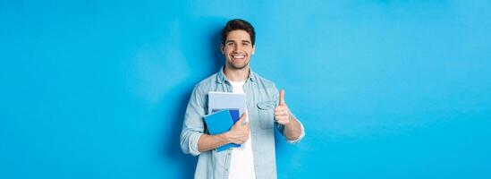 Young man student with notebooks, showing thumb up in approval, smiling satisfied, blue studio background photo