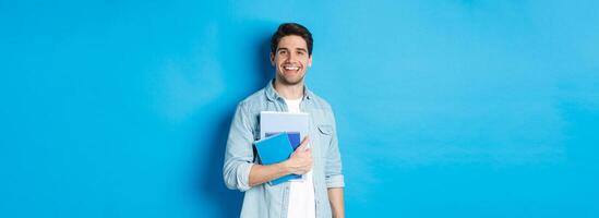 Smiling man studying, holding notebooks and looking happy, standing over blue background photo