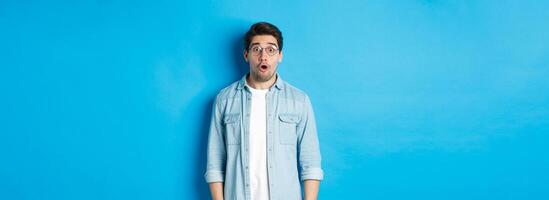 Surprised man in glasses looking at promo offer, saying wow amazed, standing over blue background photo