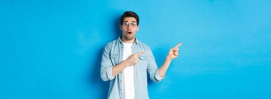 Surprised man in glasses pointing fingers left at copy space, asking question about product, looking intrigued, standing over blue background photo