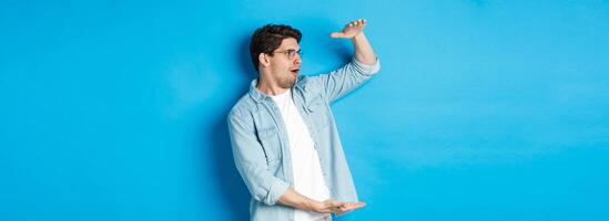 Excited handsome man showing big size object and looking amazed, standing over blue background photo