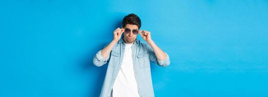 Confident macho man put on sunglasses, looking cool and sassy, standing over blue background photo