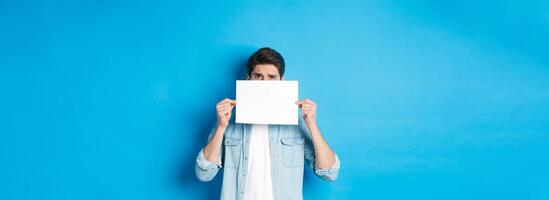 Shy and nervous guy hiding face behind blank piece of paper, peeking with worried look, showing your logo, standing over blue background photo