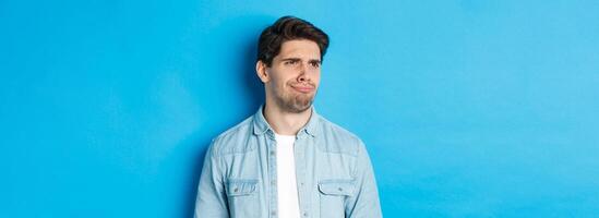 Close-up of upset and disappointed man grimacing, looking dissatisfied left, cringe from bad promo banner, standing over blue background photo
