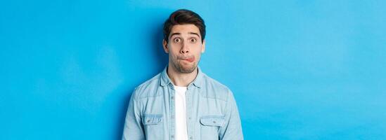 Close-up of young man making funny expressions, showing tongue and looking at camera, standing over blue background photo