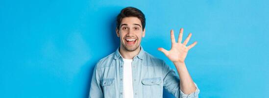 Close-up of handsome man smiling, showing fingers number five, standing over blue background photo