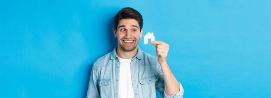 Real estate concept. Excited guy looking at small house model and smiling, renting apartment, standing over blue background photo