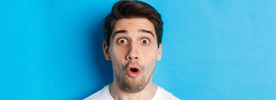 Headshot of surprised man gasping wondered, saying wow and looking amazed at camera, standing over blue background photo